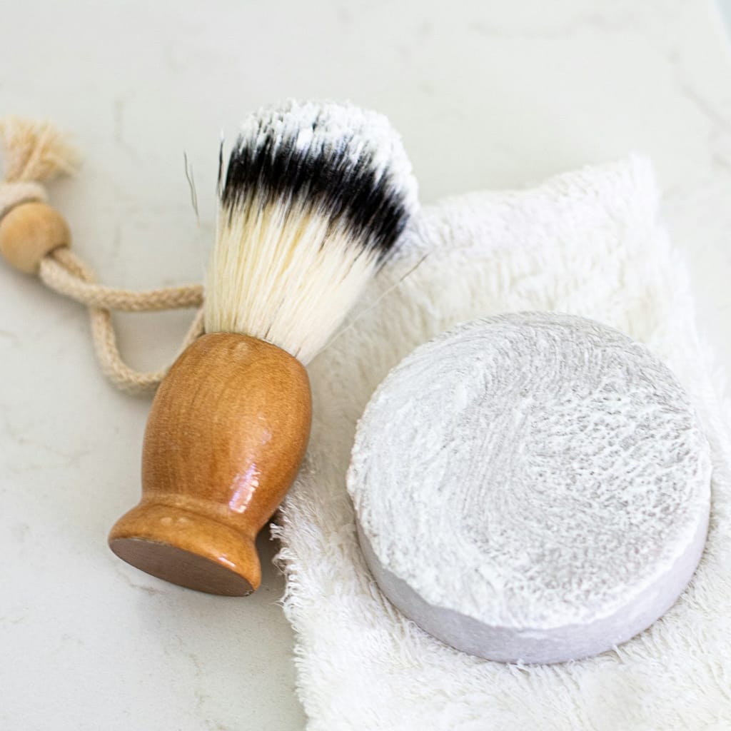 brown wooden handle shaving brush on white textile next to zero waste shaving bar